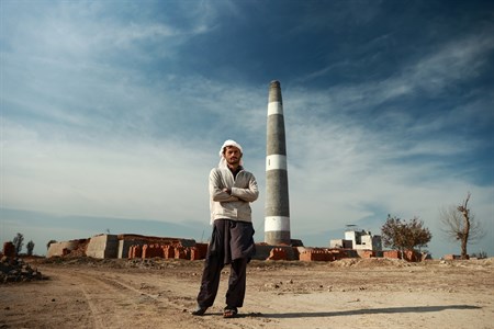 man in traditional clothes standing with folded arms with furnace in the background