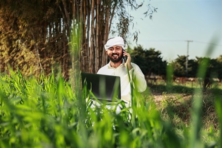 Man in traditional clothes smiling, holding a laptop while taking call on mobile phone