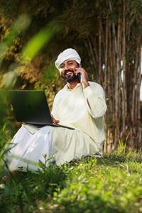man in traditional clothes smiling, holding a laptop while taking call on mobile phone