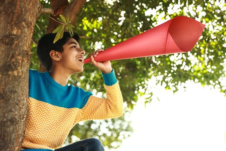 Playful kid holding a red paper speaker while sitting on tree