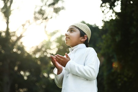 Kid praying with hands in the air wearing traditional clothes and muslim cap